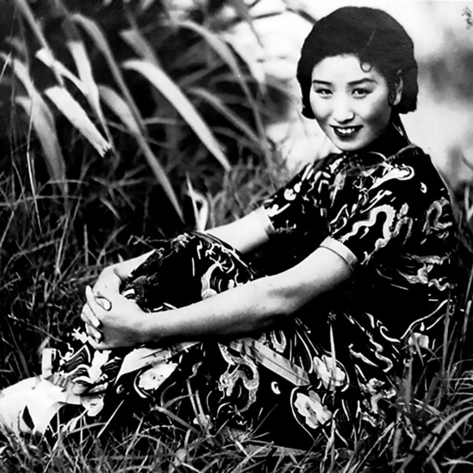 Eine Frau in einem chinesischen Kleid sitzt lächelnd im Gras, die Arme anmutig um die Beine geschlungen, schwarz-weiß, um 1940