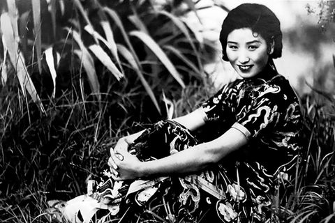 Eine Frau in einem chinesischen Kleid sitzt lächelnd im Gras, die Arme anmutig um die Beine geschlungen, schwarz-weiß, um 1940