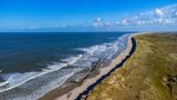 Expansive Beach on Norderney Island, Germany.  A breathtaking aerial view of the expansive beach on Norderney Island, one of the East Frisian Islands in Germany. The photograph captures the long stretch of sandy shoreline meeting the gentle waves of the North Sea, with the dune-covered landscape adding to the serene atmosphere. The vibrant blue sky and the tranquil setting make this a perfect depiction of Norderney’s natural beauty, offering an inviting glimpse of this popular destination for beachgoers and nature enthusiasts.