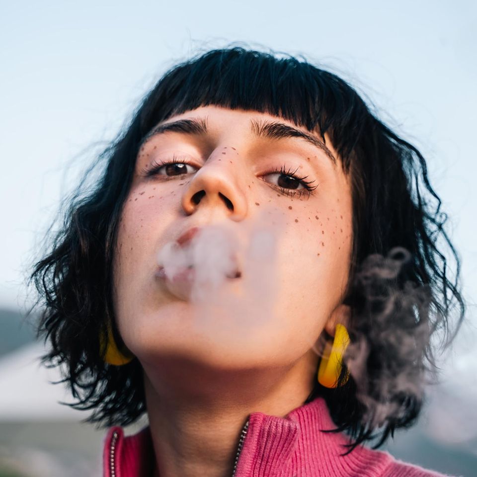 Junge Frau bläst mit halb geöffnetem Mund Rauch in Richtung Kamera