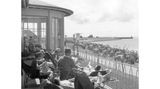 Büsum, 1957 Ein friedlicher Moment am Meer: Die Sonne wärmt die Terrasse der Lesehalle im Seebad Büsum. Aus dem Liegestuhl schweift der Blick über die Reihen von Strandkörben – dahinter liegt die Einfahrt zum Fischereihafen.