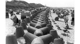 Wenningstedt, 1984 Ein massiver Betongürtel soll auf Sylt den Badespaß bewahren: Die mächtige Struktur verläuft mitten durch das Strandidyll, um bei Hochwasser das Abtragen der Küste zu verhindern. Die vierarmigen Betonklötze (auch Tetrapoden genannt) wurden auf der Insel ab den 1960er-Jahren verbaut. Doch ihr Nutzen blieb umstritten – sie verrutschten bei Sturmfluten, förderten stellenweise den Sandverlust und störten das Landschaftsbild. Umweltfragen und Kritik an ihrer Ästhetik machten sie bald zum Symbol für gescheiterten Küstenschutz. Viele Tetrapoden wurden inzwischen abgeräumt und lagern heute ungenutzt auf der Insel. 2023 erhielten sie neues Leben: Unbekannte sprühten Augen auf die grauen Klötze – aus Küstenschutz wurde Kunst.