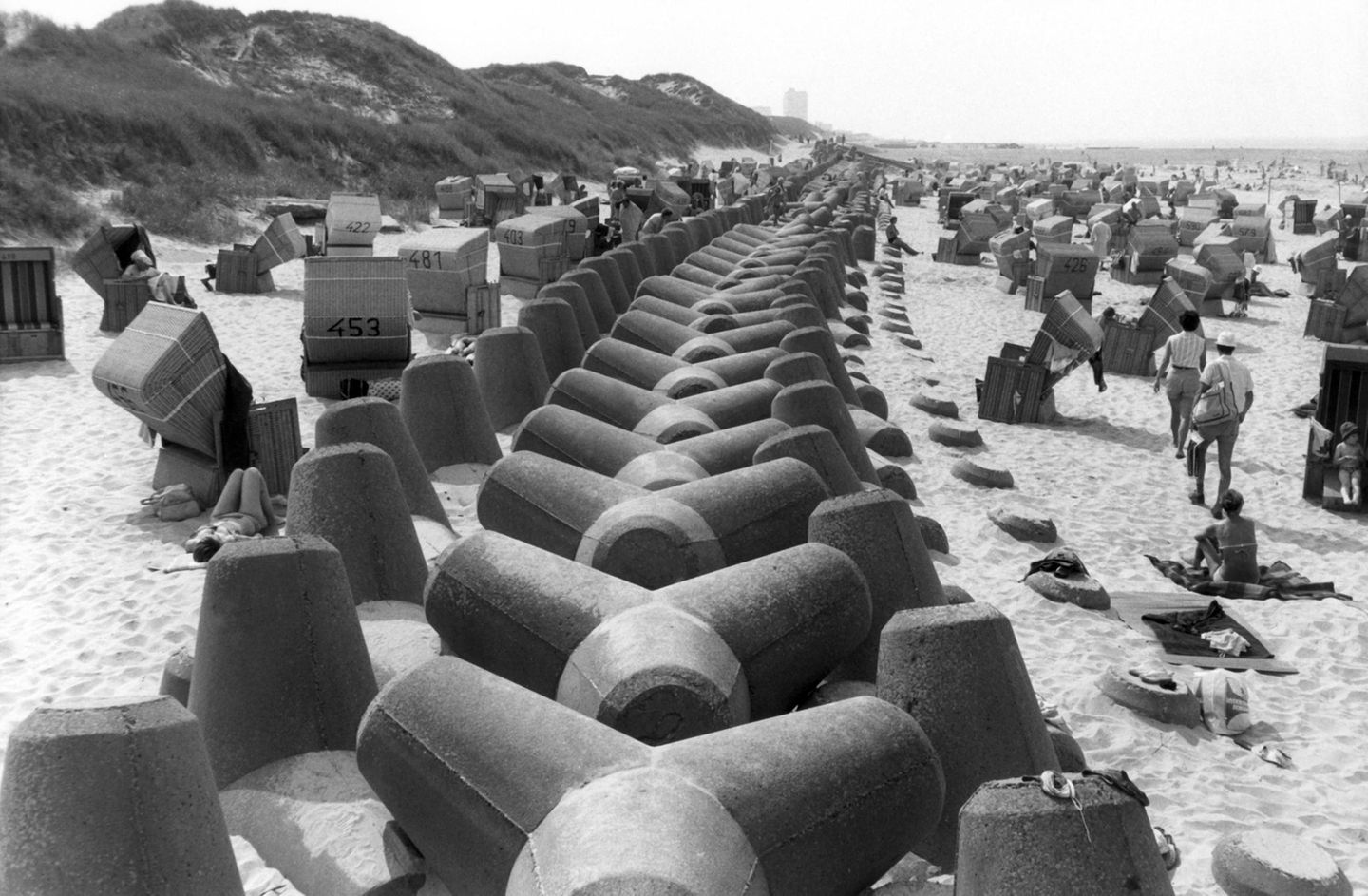 Wenningstedt, 1984 Ein massiver Betongürtel soll auf Sylt den Badespaß bewahren: Die mächtige Struktur verläuft mitten durch das Strandidyll, um bei Hochwasser das Abtragen der Küste zu verhindern. Die vierarmigen Betonklötze (auch Tetrapoden genannt) wurden auf der Insel ab den 1960er-Jahren verbaut. Doch ihr Nutzen blieb umstritten – sie verrutschten bei Sturmfluten, förderten stellenweise den Sandverlust und störten das Landschaftsbild. Umweltfragen und Kritik an ihrer Ästhetik machten sie bald zum Symbol für gescheiterten Küstenschutz. Viele Tetrapoden wurden inzwischen abgeräumt und lagern heute ungenutzt auf der Insel. 2023 erhielten sie neues Leben: Unbekannte sprühten Augen auf die grauen Klötze – aus Küstenschutz wurde Kunst.