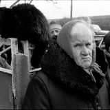 Ältere Person guckt die Betrachtenden direkt an. Links neben ihr auf einem Tor sitzt eine schwarze Katze.