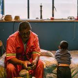 Person sitzt in roter Arbeiterkleidung auf einem Bett. neben ihm sitzt ein Junge, der ihm den Rücken zugekehrt hat.