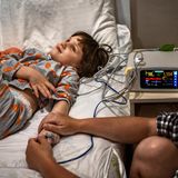 Junges Mädchen liegt auf einem Krankenhausbett und guckt an die Zimmerdecke, während erwachsene person ihre Hand hält.