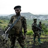 Soldaten stehen in Millitäruniform mit Gewehren auf einem Hügel. Im Hintergrund sieht man eine Stadt.