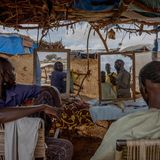 Zwei Personen sitzen mit dem Rücken zu den Betrachtenden und gucken in Spiegel, während sie rasiert werden.