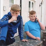 Zwei Jungen stehen vor einem Haus. Einer pustet in einen Platikbecher. Einer hält ein Smartphone an sein Ohr.