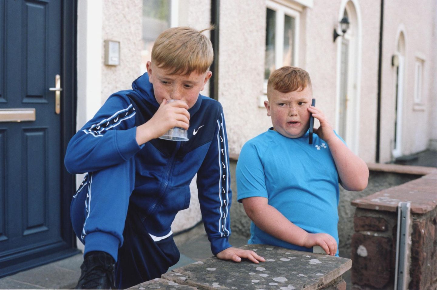 Zwei Jungen stehen vor einem Haus. Einer pustet in einen Platikbecher. Einer hält ein Smartphone an sein Ohr.