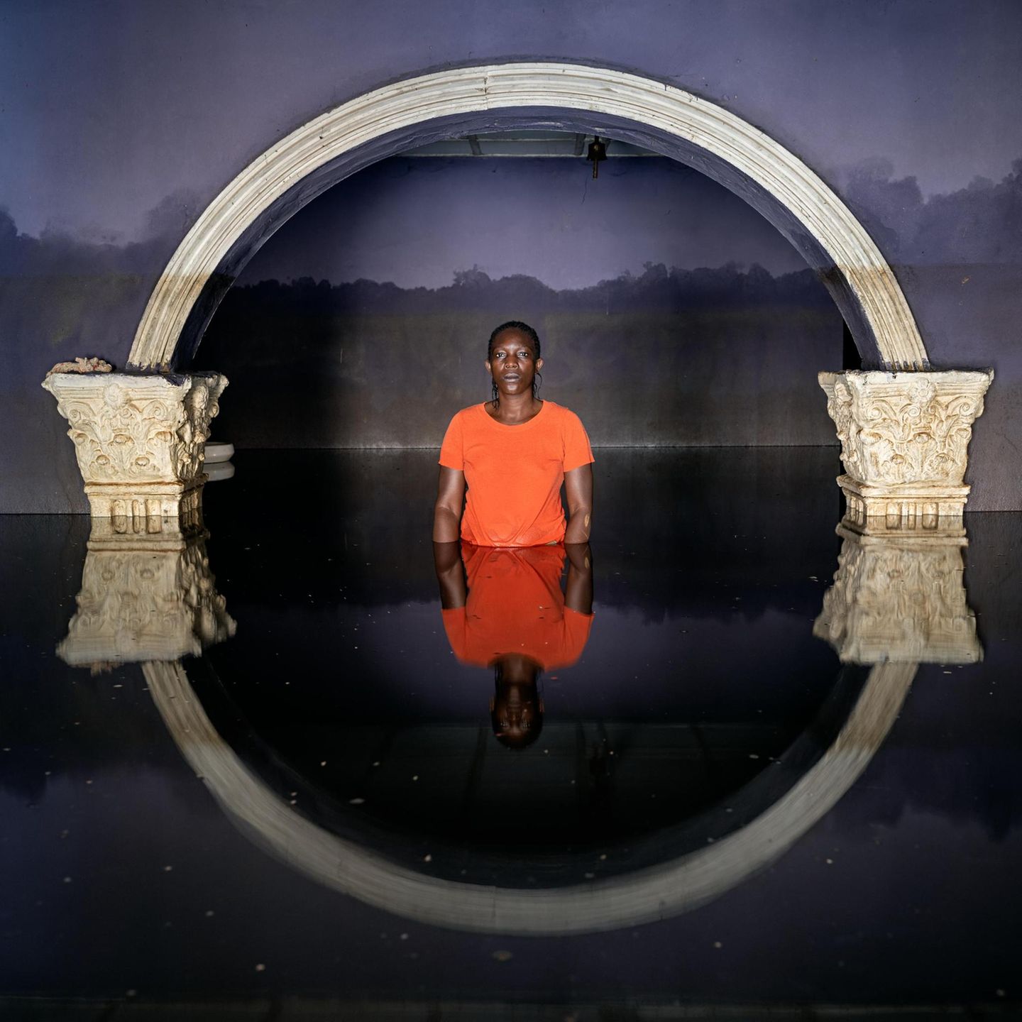 Eine Person in roter Kleidung steht bis zur Hüfte in Wasser. Über ihr ein Torbogen, der sich in der Wasseroberfläche spiegelt.