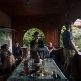 Eine Gruppe Menschen sitzen um einen Tisch herum auf einer Veranda. Eine Person im Hintergrund hält ein Gewehr bereit.