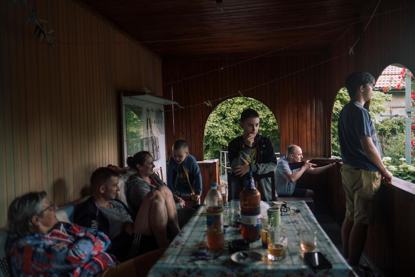 Nostalgie Eine Gruppe Menschen sitzen um einen Tisch herum auf einer Veranda. Eine Person im Hintergrund hält ein Gewehr bereit.