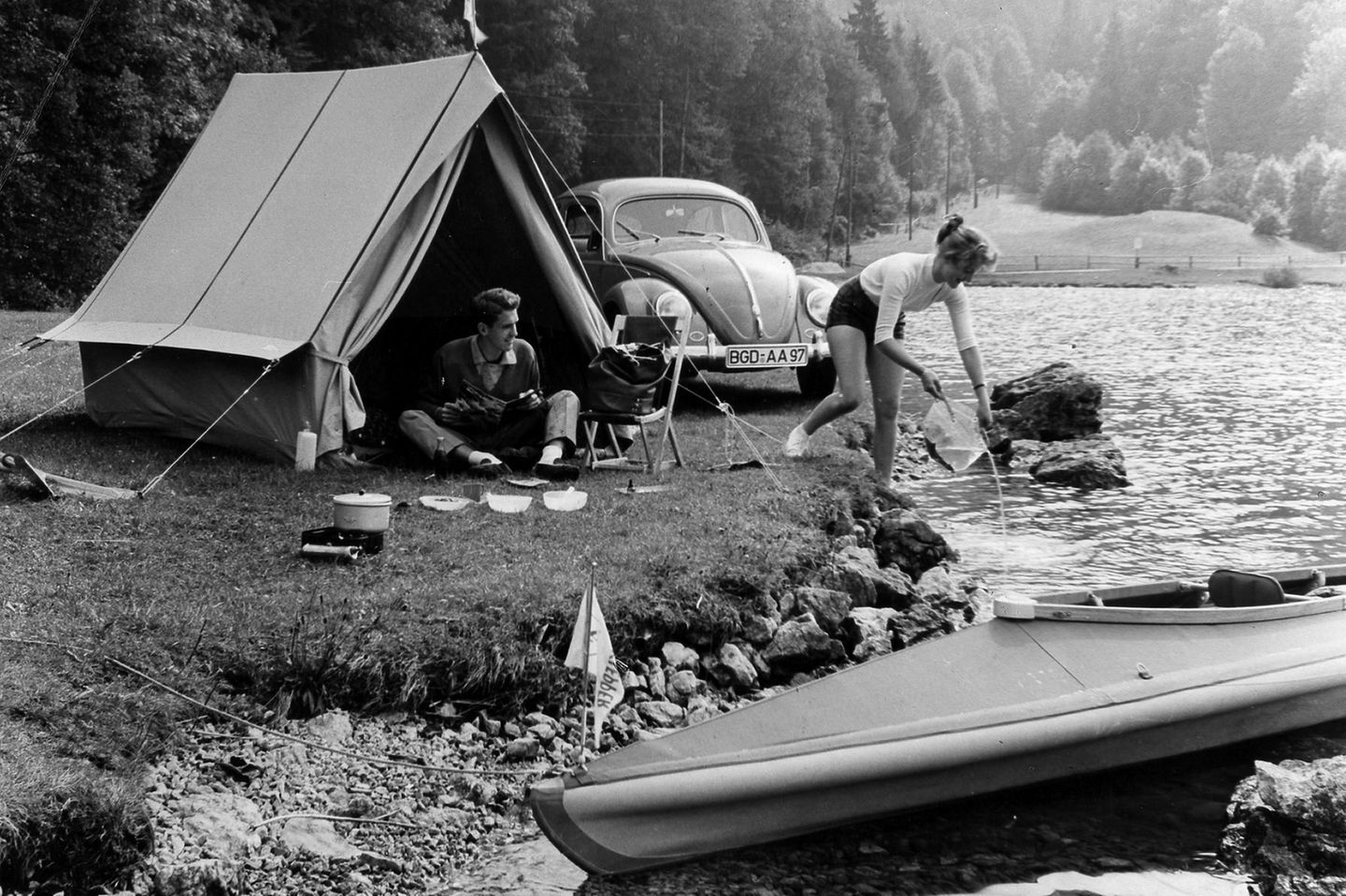 Junges Paar zeltet mit einem Käfer an einem Fluss, 1956