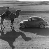 Ein Kamelreiter steht vor einem Käfer in der Wüste