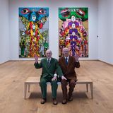 Gilbert und George sitzen auf einer Bank in ihrer Ausstellung und winken, 2025