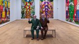 Gilbert und George sitzen auf einer Bank in ihrer Ausstellung und winken, 2025