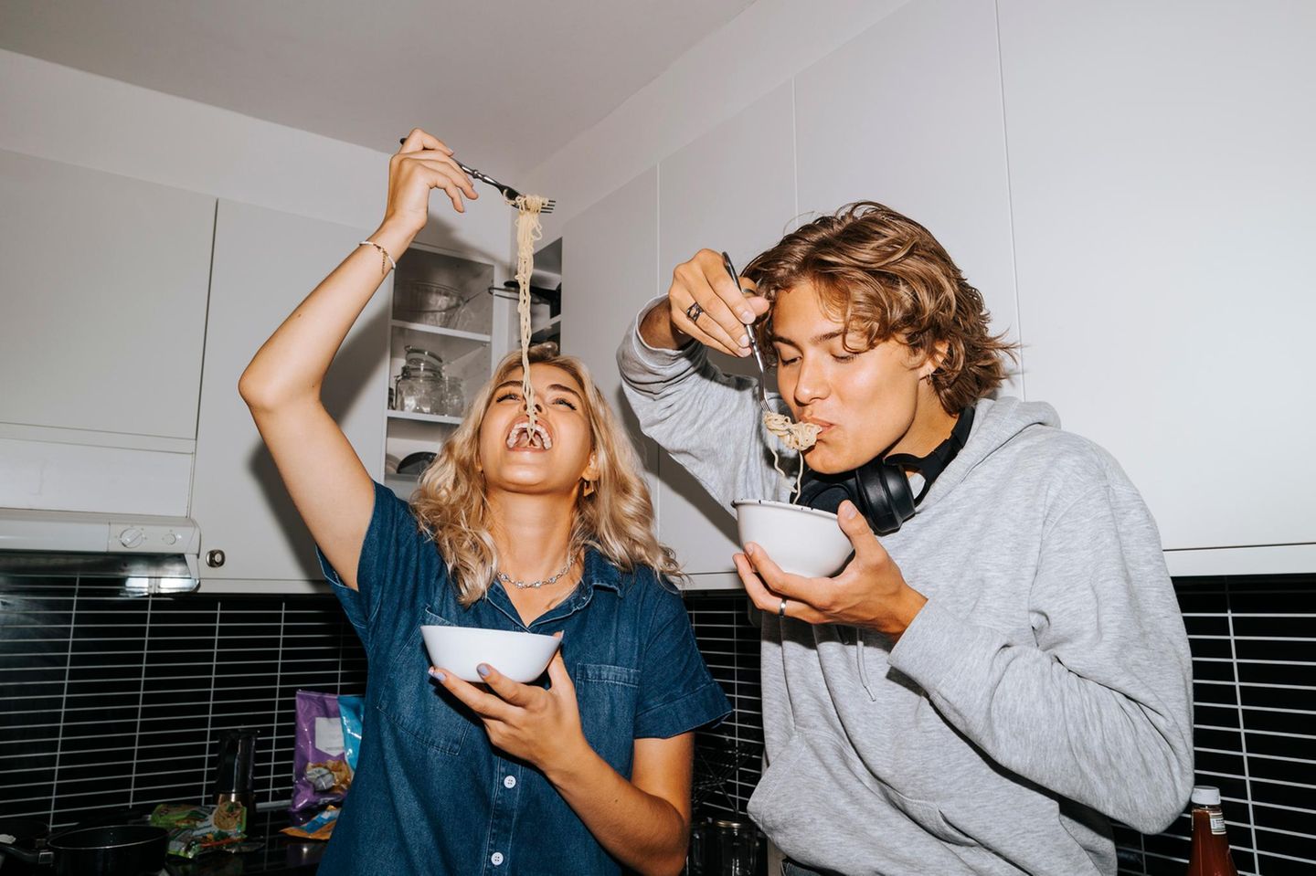 Junge Frau und Mann stehen nebeinander in der Küche und essen spaßhaft gemeinsam Spaghetti aus einer kleinen Schüssel.