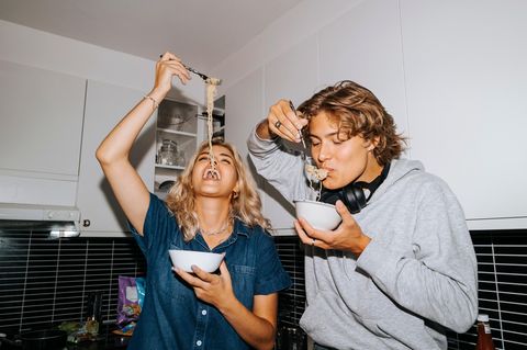 Junge Frau und Mann stehen nebeinander in der Küche und essen spaßhaft gemeinsam Spaghetti aus einer kleinen Schüssel.