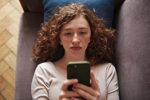 Mädchen mit roten Locken liegt auf dem Sofa und scrollt in ihrem Smartphone