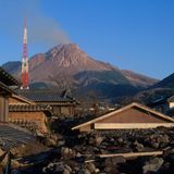 Von Geröll und Staub bedeckte Häuser, im Hintergrund der Unzen, aus dem leichter Rauch empor steigt