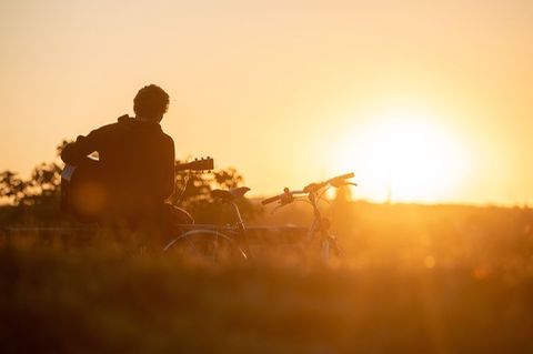 Mann spielt bei Sonnenuntergang Gitarre in der Natur. Neben ihm steht ein Fahrrad