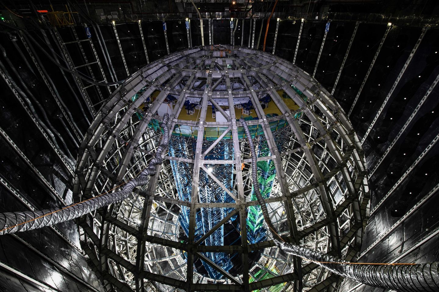 Wen soll diese Kugel aufstöbern? Geisterteilchen. Neutrinos sind die leichtesten und zugleich am schwersten detektierbaren Teilchen des Kosmos. In jeder Sekunde fliegen Zigmilliarden durch einen menschlichen Körper, ohne dass wir es bemerken. Diese 35 Meter hohe kugelförmige Stahlkonstruktion, von chinesischen Forschenden kürzlich fertiggestellt, soll sie aufspüren und ihnen ihre Geheimnisse entlocken. Umgeben mit einem Panzer aus Plexiglas fasst die Kugel 20.000 Tonnen einer Flüssigkeit, die aufleuchtet, wenn Neutrinos sie durchqueren. Um dem Innendruck der Flüssigkeit auf die Außenfläche etwas entgegenzusetzen, schwebt die Kugel in einem riesigen Wasserbecken, 44 Meter tief, angefüllt mit 60.000 Tonnen hochreinem Wasser. Und als wäre das nicht schon megalomanisch genug, befindet sich die gesamte Konstruktion 700 Meter unter der Erde, um sie vor äußeren Störungen zu schützen. Solch ein Aufwand wäre wohl übertrieben, wäre das Neutrino nicht das mysteriöseste und am wenigsten verstandene Elementarteilchen. Forschende können viele seiner Eigenschaften nicht erklären: Es besitzt eine (winzige) Masse, auch wenn es das nicht sollte; es kann sich nur in eine Richtung drehen und ein einzelnes wandelt sich unentwegt in verschiedene Arten von Neutrinos um. Mit der Kugel wollen Forschende dieses ungewöhnliche Verhalten genauer beobachten und verstehen, in der Erwartung und Hoffnung, etwas fundamental Neues über das Universum zu lernen. KW 36