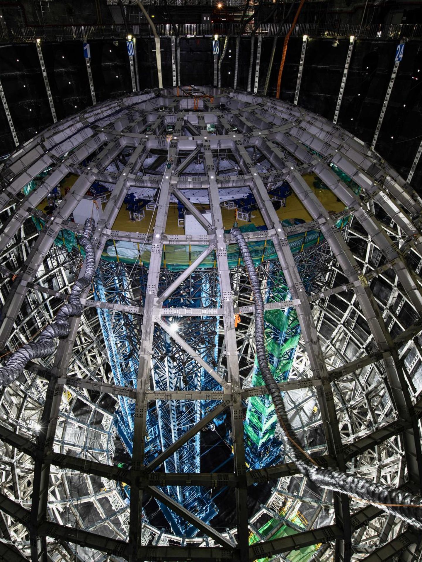 Geisterteilchen. Neutrinos sind die leichtesten und zugleich am schwersten detektierbaren Teilchen des Kosmos. In jeder Sekunde fliegen Zigmilliarden durch einen menschlichen Körper, ohne dass wir es bemerken. Diese 35 Meter hohe kugelförmige Stahlkonstruktion, von chinesischen Forschenden kürzlich fertiggestellt, soll sie aufspüren und ihnen ihre Geheimnisse entlocken.   Umgeben mit einem Panzer aus Plexiglas fasst die Kugel 20.000 Tonnen einer Flüssigkeit, die aufleuchtet, wenn Neutrinos sie durchqueren. Um dem Innendruck der Flüssigkeit auf die Außenfläche etwas entgegenzusetzen, schwebt die Kugel in einem riesigen Wasserbecken, 44 Meter tief, angefüllt mit 60.000 Tonnen hochreinem Wasser. Und als wäre das nicht schon megalomanisch genug, befindet sich die gesamte Konstruktion 700 Meter unter der Erde, um sie vor äußeren Störungen zu schützen.   Solch ein Aufwand wäre wohl übertrieben, wäre das Neutrino nicht das mysteriöseste und am wenigsten verstandene Elementarteilchen. Forschende können viele seiner Eigenschaften nicht erklären: Es besitzt eine (winzige) Masse, auch wenn es das nicht sollte; es kann sich nur in eine Richtung drehen und ein einzelnes wandelt sich unentwegt in verschiedene Arten von Neutrinos um. Mit der Kugel wollen Forschende dieses ungewöhnliche Verhalten genauer beobachten und verstehen, in der Erwartung und Hoffnung, etwas fundamental Neues über das Universum zu lernen.  KW 36