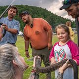 Ein Kind hält eine Klapperschlange in der Hand, um sie herum erwachsene Personen