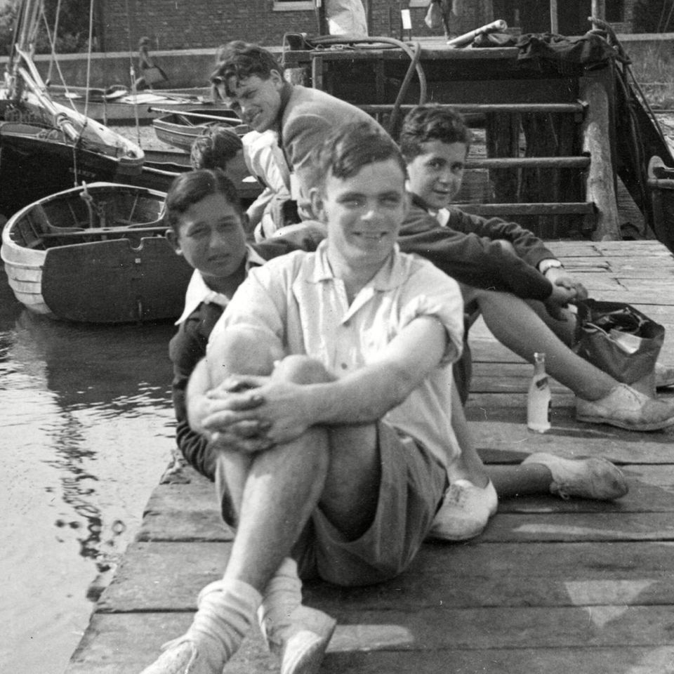 Alan Turing sitze auf einem Steg und blickt in die Kamera. Hinter ihm sitzen weitere junge Männer