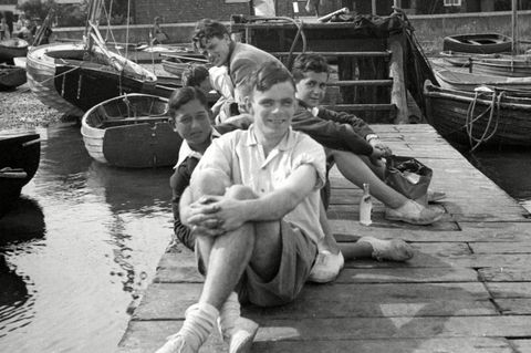Alan Turing sitze auf einem Steg und blickt in die Kamera. Hinter ihm sitzen weitere junge Männer