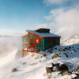 Versteckt auf einem Granitrücken, tief im Herzen von British Columbias Gebirgslandschaft, wird schon der Weg zur Jim Haberl Hut zur Herausforderung – eine anstrengende Expedition durch felsiges, hochalpines Gelände. Auf über 2.000 Metern schließlich wartet diese kompakte, solide und zweckmäßige Unterkunft. Mit ihrem seegrünen Farbton und den kräftigen, orangefarbenen Details ist die funktionale Hütte ein hübscher Farbklecks in der kargen Landschaft. Das matt schimmernde Wellblechdach verändert je nach Wetter sein Aussehen – es leuchtet im Sonnenlicht, bei bewölktem Himmel erscheint es eher grau. 
