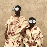 Zwei junge Menschen posieren vor einer rauen Wand, in reich bestickten Gewändern aus Aso Oke – jenem handgewebten Stoff aus Nigeria, der traditionell bei Zeremonien getragen wird. Ihre Gesichter sind größtenteils von futuristischen Brillen aus recycelten VHS-Kassetten verdeckt – eine witzige und prägnante Metapher für die Verwandlung veralteter Technik in ein neues Ritualobjekt und Werkzeug des kulturellen Ausdrucks.