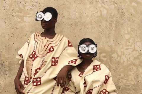 Zwei junge Menschen posieren vor einer rauen Wand, in reich bestickten Gewändern aus Aso Oke – jenem handgewebten Stoff aus Nigeria, der traditionell bei Zeremonien getragen wird. Ihre Gesichter sind größtenteils von futuristischen Brillen aus recycelten VHS-Kassetten verdeckt – eine witzige und prägnante Metapher für die Verwandlung veralteter Technik in ein neues Ritualobjekt und Werkzeug des kulturellen Ausdrucks.
