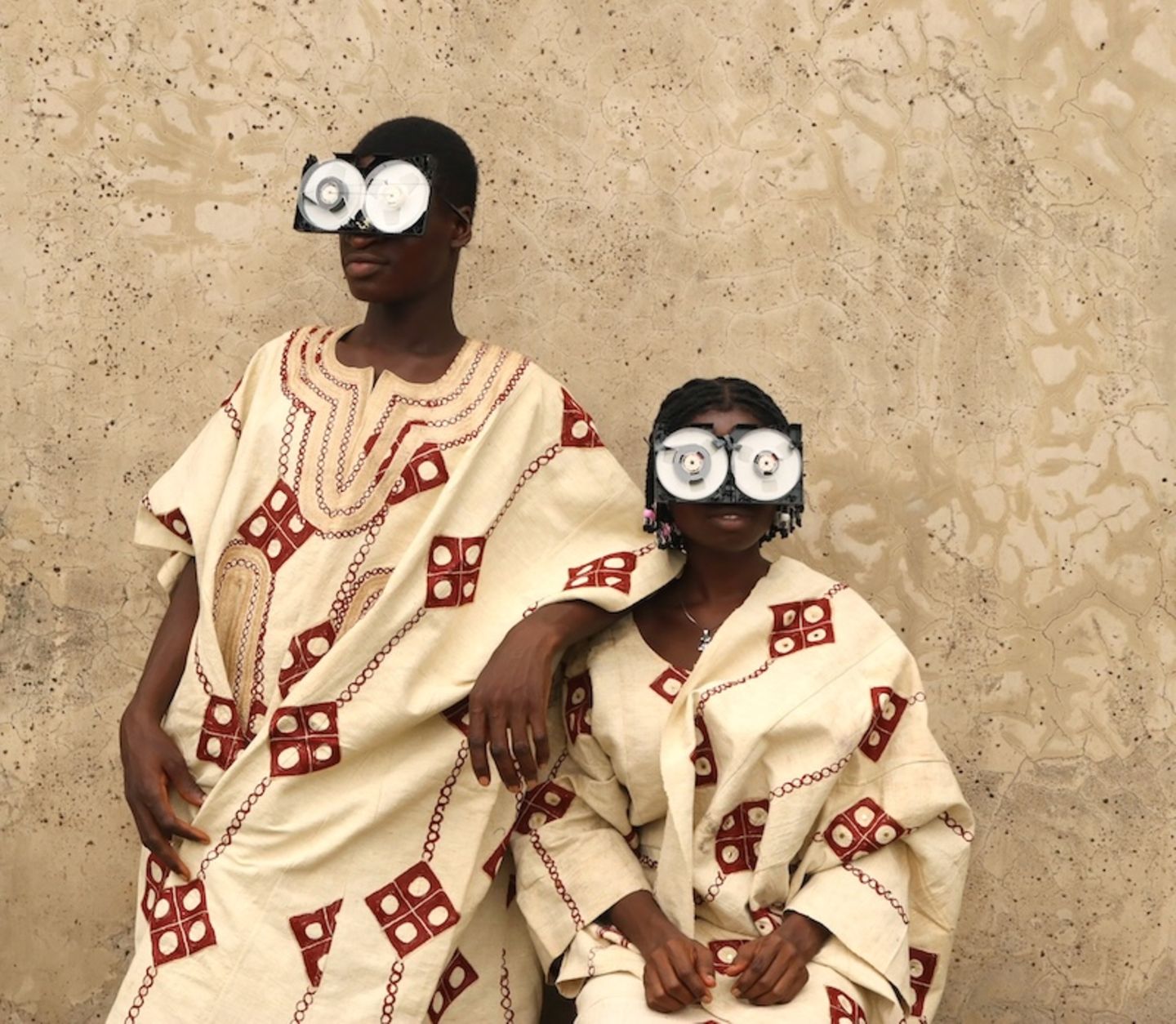 Zwei junge Menschen posieren vor einer rauen Wand, in reich bestickten Gewändern aus Aso Oke – jenem handgewebten Stoff aus Nigeria, der traditionell bei Zeremonien getragen wird. Ihre Gesichter sind größtenteils von futuristischen Brillen aus recycelten VHS-Kassetten verdeckt – eine witzige und prägnante Metapher für die Verwandlung veralteter Technik in ein neues Ritualobjekt und Werkzeug des kulturellen Ausdrucks.