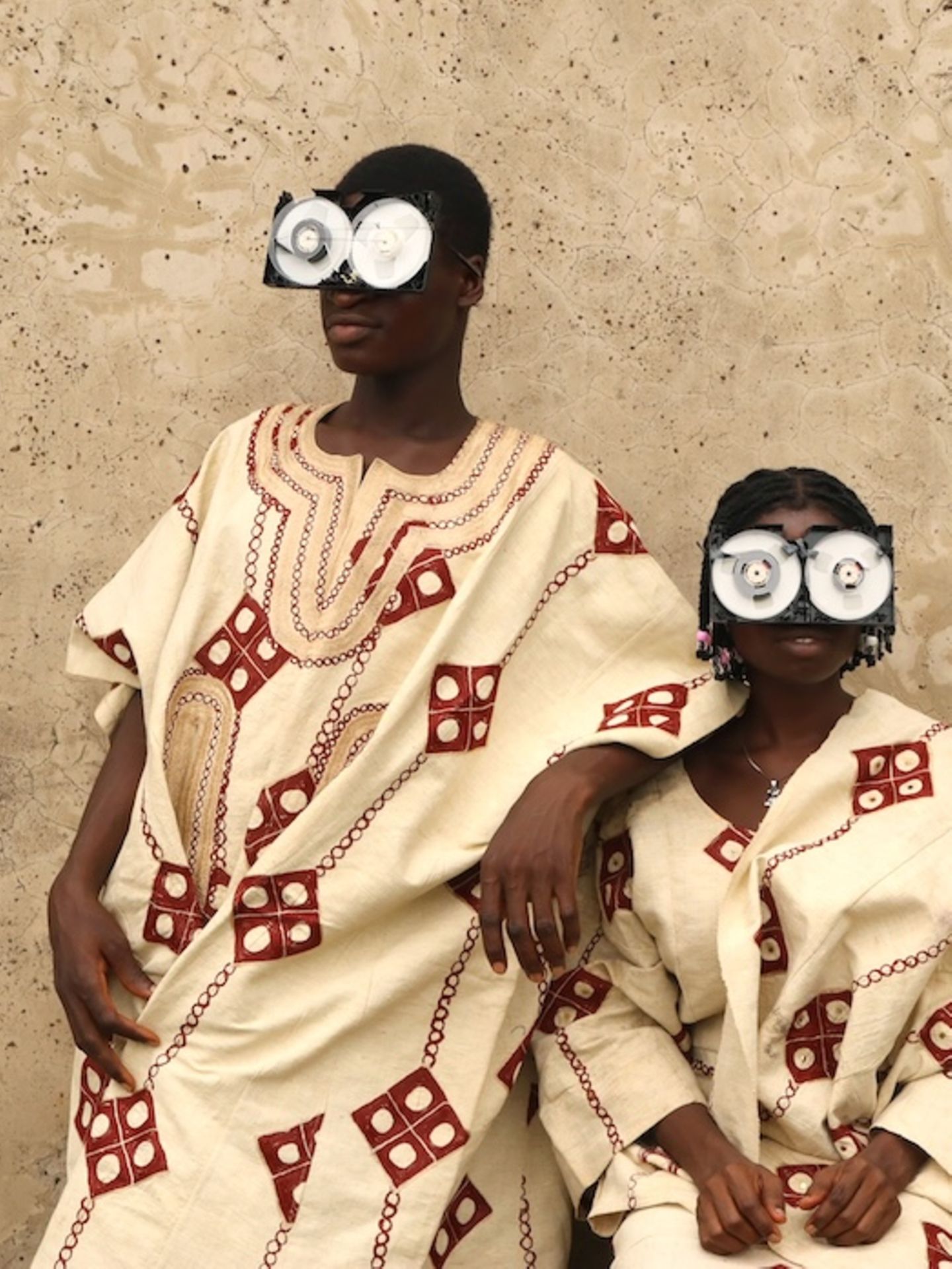 Zwei junge Menschen posieren vor einer rauen Wand, in reich bestickten Gewändern aus Aso Oke – jenem handgewebten Stoff aus Nigeria, der traditionell bei Zeremonien getragen wird. Ihre Gesichter sind größtenteils von futuristischen Brillen aus recycelten VHS-Kassetten verdeckt – eine witzige und prägnante Metapher für die Verwandlung veralteter Technik in ein neues Ritualobjekt und Werkzeug des kulturellen Ausdrucks.