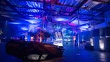 Blau beleuchete Ausstellungshalle mit beschädigtem Auto und Videoinstallationen