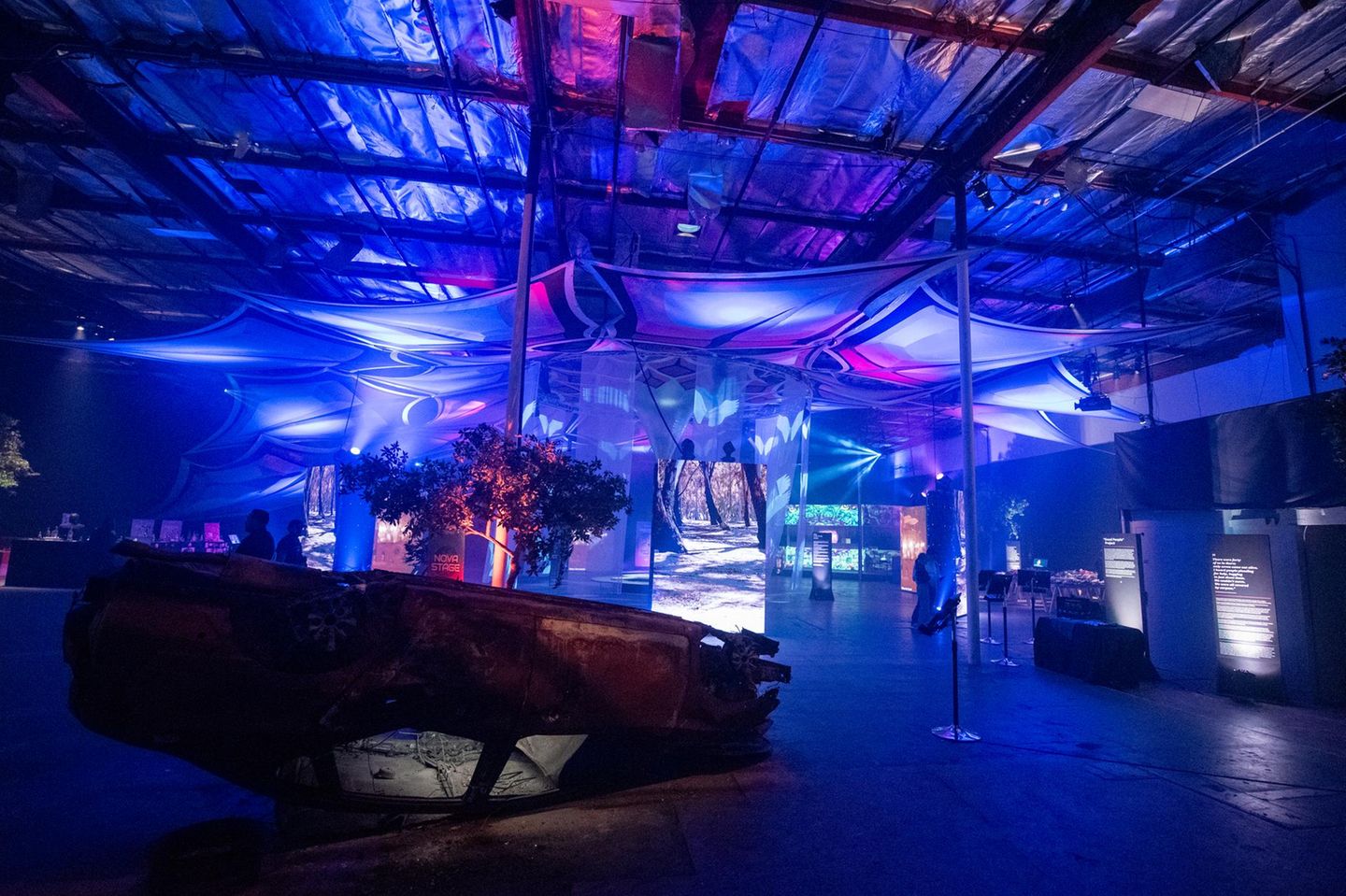 Blau beleuchete Ausstellungshalle mit beschädigtem Auto und Videoinstallationen