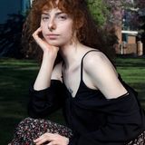 Eine junge Person mit roten lockigen Haaren sitzt auf einer Wiese und blickt in die Kamera