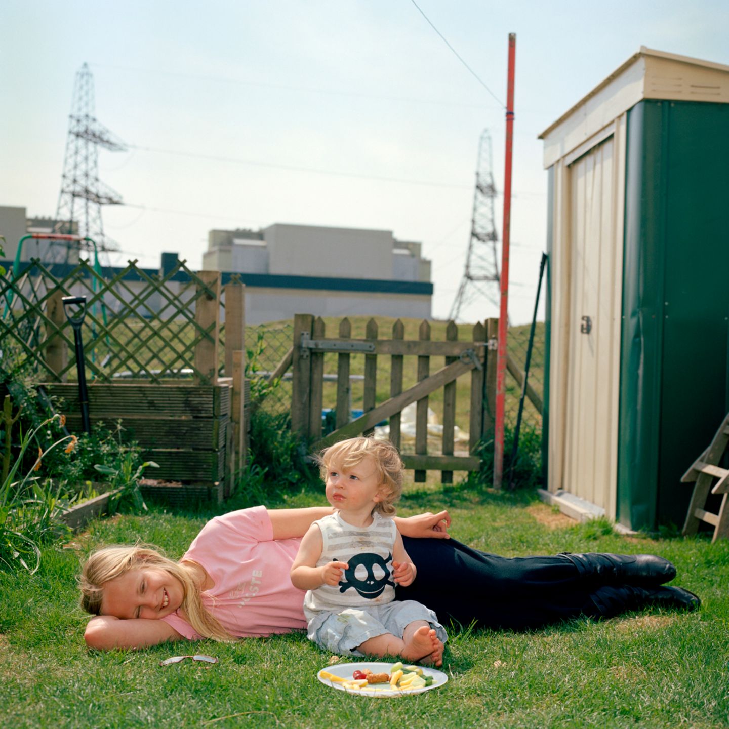 Ein Kind liegt im Garten, während ein Kleinkind vor ihr auf dem Rasen kniet. Vor ihnen ein Teller mit Essen