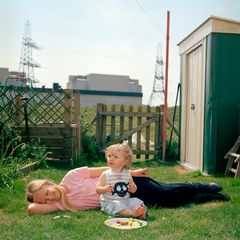 Ein Kind liegt im Garten, während ein Kleinkind vor ihr auf dem Rasen kniet. Vor ihnen ein Teller mit Essen