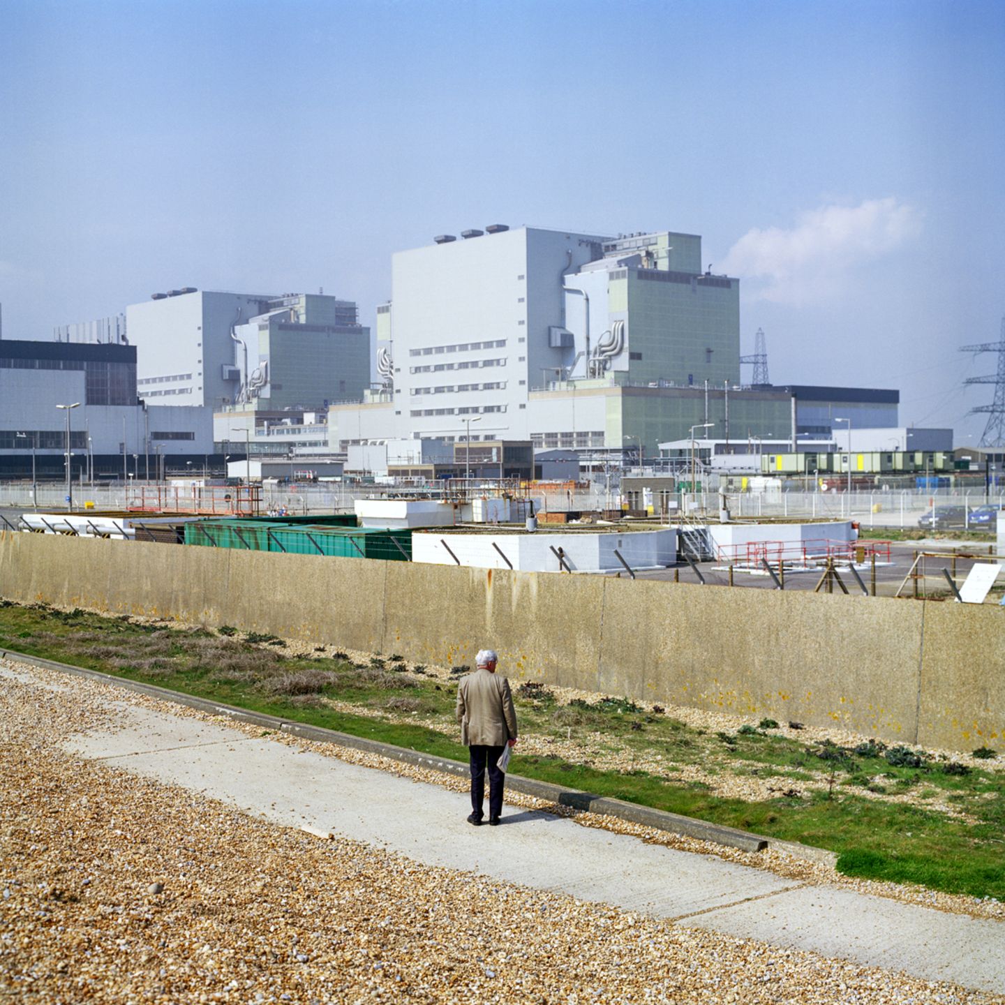 Eine alte Person steht vor einem Zaun, hinter dem sich eine riesige Anlage befindet