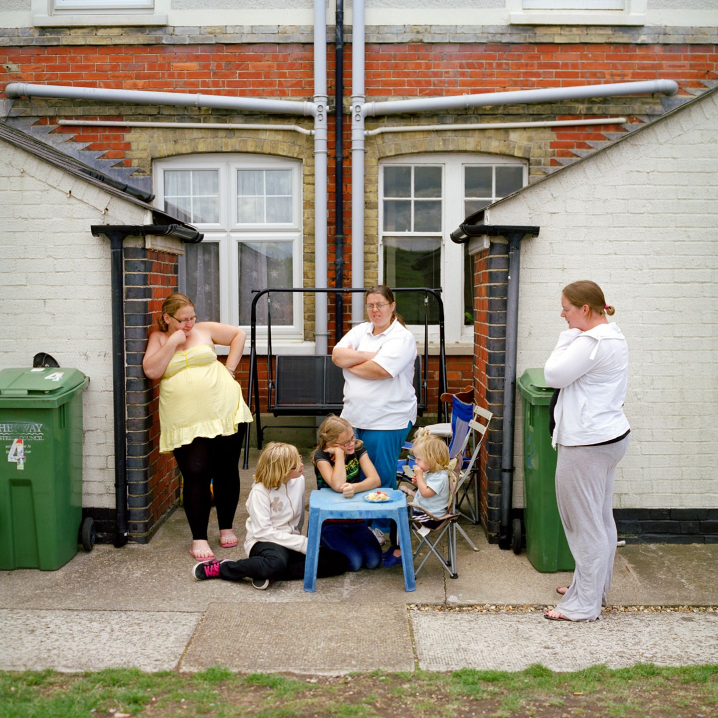 Drei erwachsene Personen, die vor einem Haus stehen. Drei Kinder sitzen davor an einem Tisch