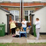 Drei erwachsene Personen, die vor einem Haus stehen. Drei Kinder sitzen davor an einem Tisch