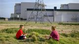 Zwei Kinder sitzen auf einer Wiese während im Hintergrund ein Strommast und eine Industrieanlage steht