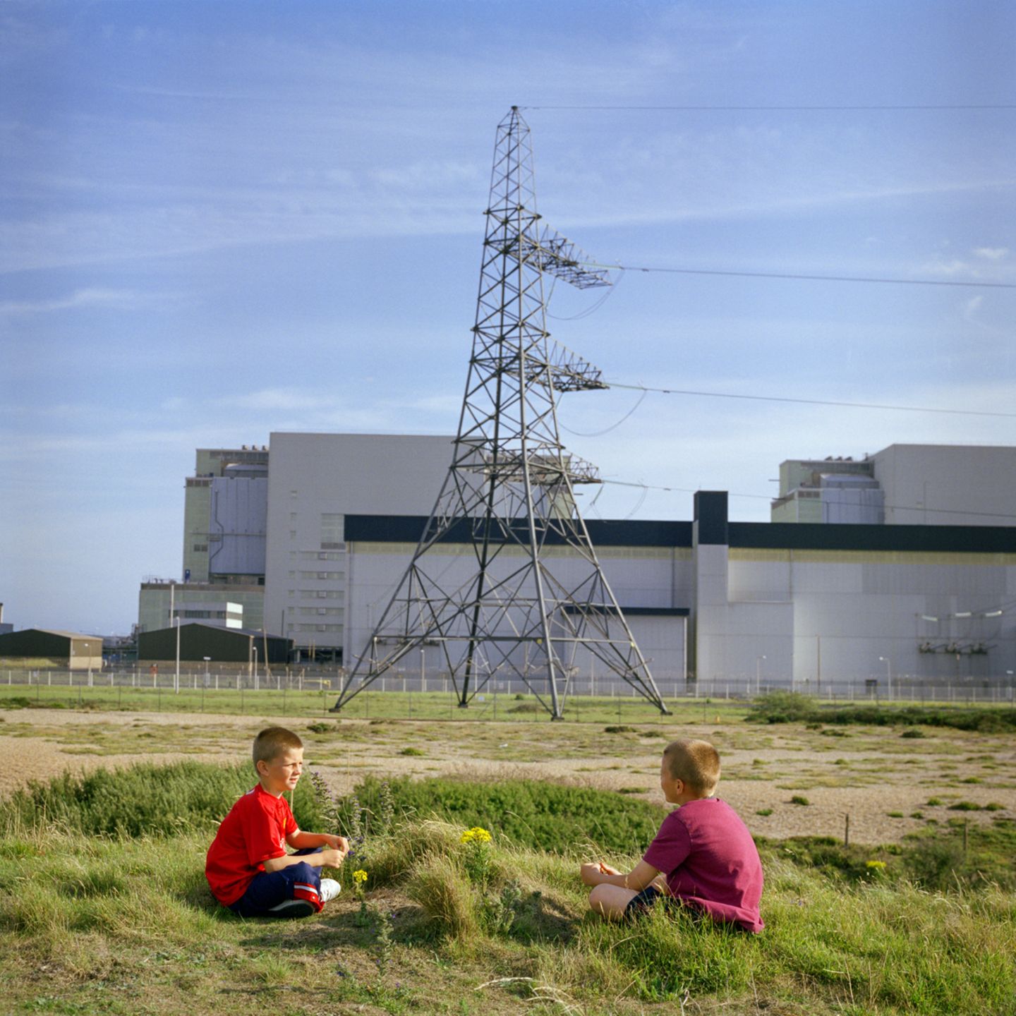 Zwei Kinder sitzen auf einer Wiese während im Hintergrund ein Strommast und eine Industrieanlage steht