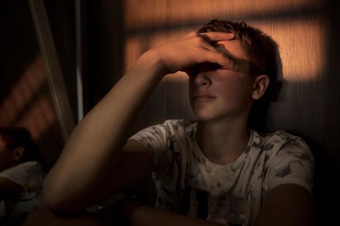 Jugendlicher lehnt in abgedunkelten Zimmer an der Wand und hält sich ein Hand vor sein Gesicht.