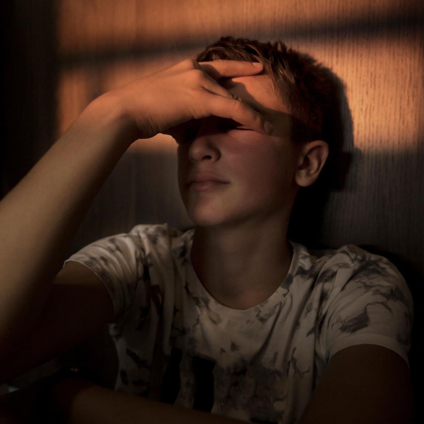 Jugendlicher lehnt in abgedunkelten Zimmer an der Wand und hält sich ein Hand vor sein Gesicht.