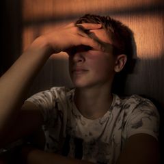 Jugendlicher lehnt in abgedunkelten Zimmer an der Wand und hält sich ein Hand vor sein Gesicht.