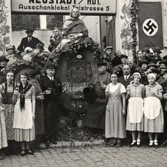 Vor einem großen Weinfass stehen Frauen in Schürzen und Männer mit gefüllten Weingläsern. Daneben hängt eine Hakenkreuzfahne.